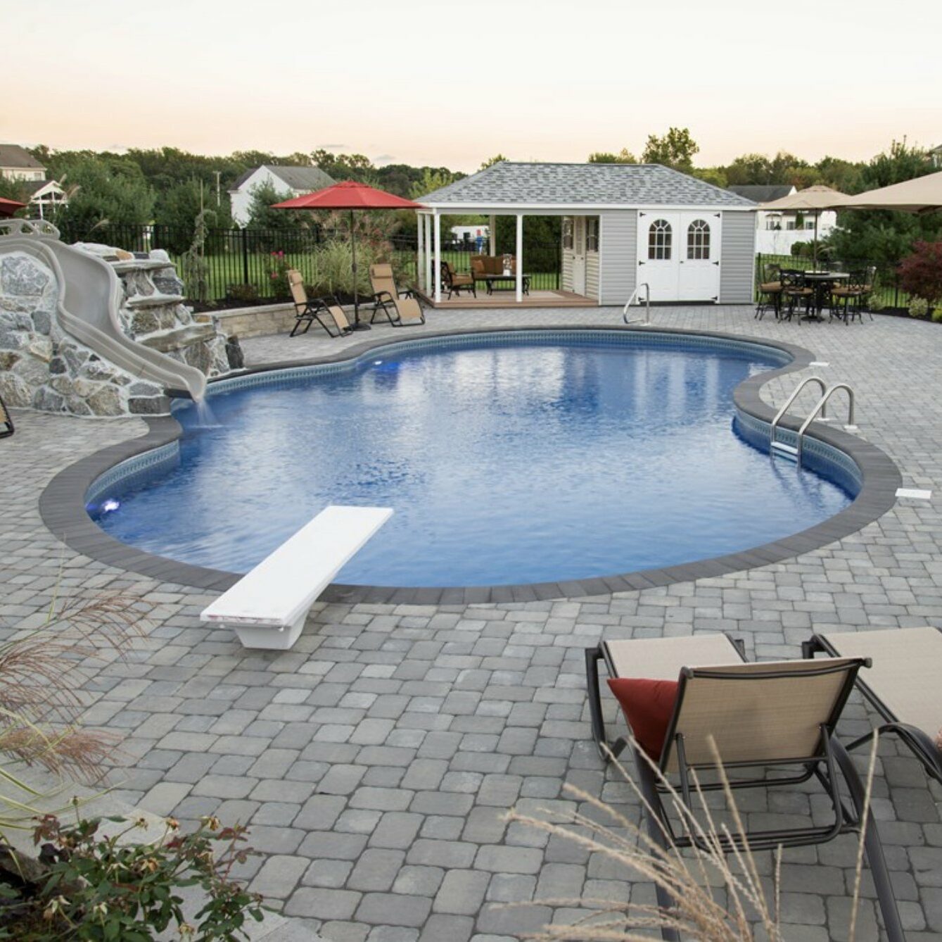 Backyard fiberglass leisure pool with a diving board, slide, and poolside lounging chairs surrounded by pavers.