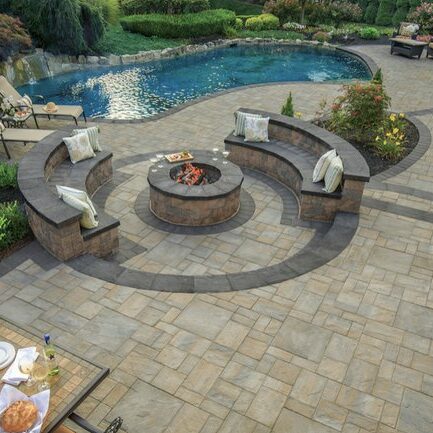 Elegant paver patio with built-in curved seating, a central fire pit, and a poolside view.