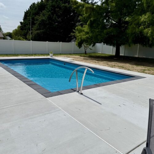 Luxury in-ground fiberglass swimming pool with a modern concrete patio and dark stone coping, installed by Coastal Outdoor Living.