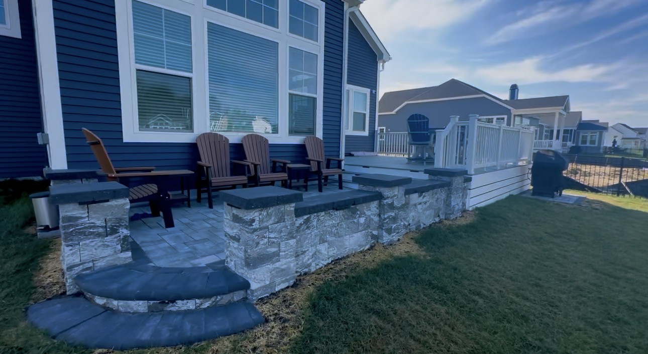 A multilevel custom deck & stone paver patio featuring a grey stone retaining wall, integrated steps, and wooden lounge chairs in a residential backyard.